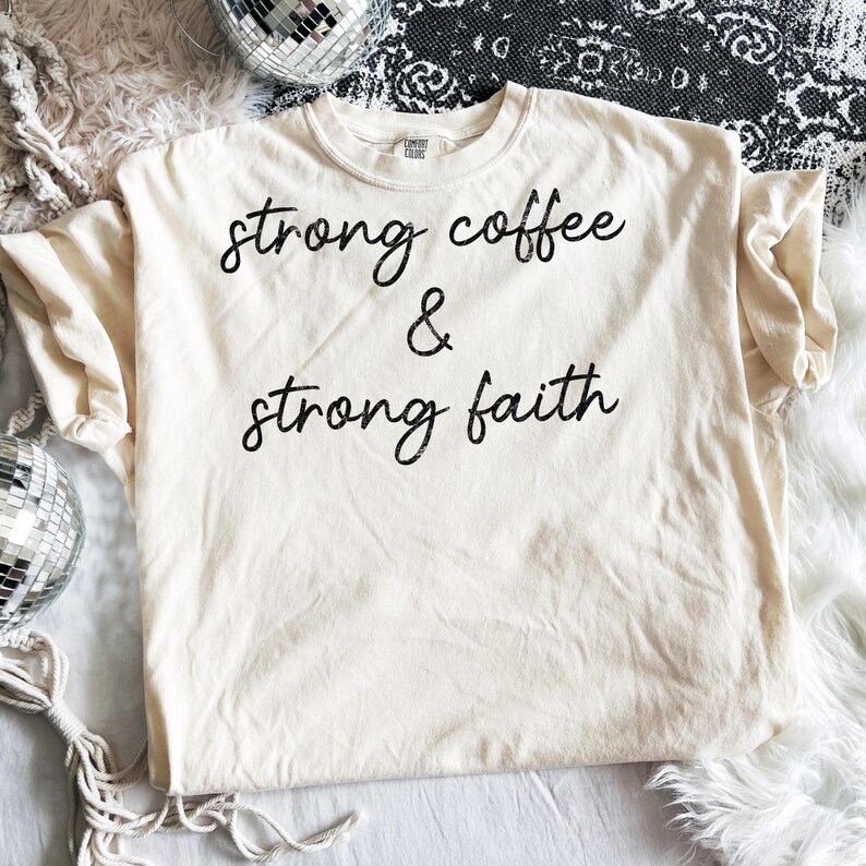 May include: Cream-colored t-shirt with the text "strong coffee & strong faith" in black script. The shirt is made of a soft material and is laid flat on a white surface. The shirt is a Comfort Colors brand.