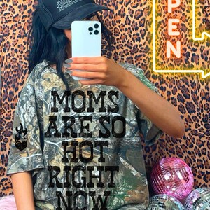 May include: A camouflage print t-shirt with the text "MOMS ARE SO HOT RIGHT NOW" in black block letters. The image also features a black trucker hat with white stitching and a neon sign that says "OPEN".