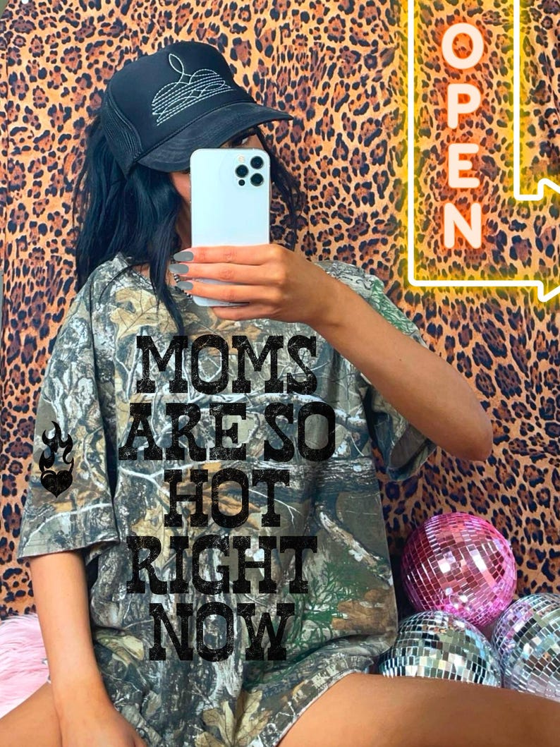 May include: A camouflage print t-shirt with the text "MOMS ARE SO HOT RIGHT NOW" in black block letters. The image also features a black trucker hat with white stitching and a neon sign that says "OPEN".