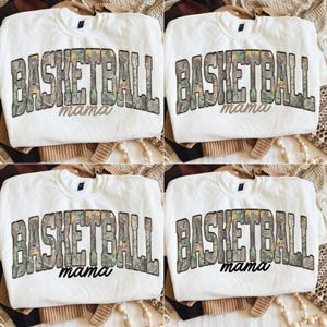 May include: Four white sweatshirts featuring the text "BASKETBALL mama" in a camouflage pattern. The sweatshirts are displayed on a neutral background. The crew neck sweatshirts are designed with a focus on the text.