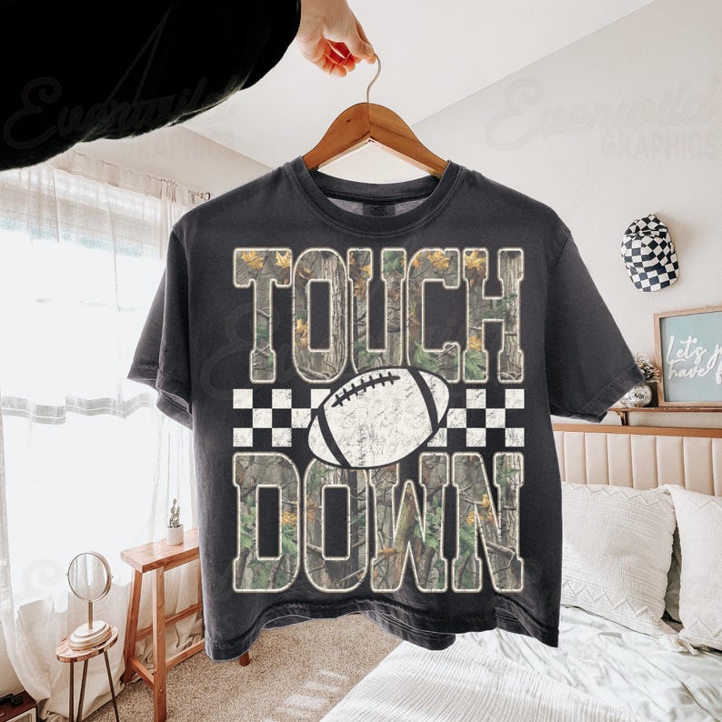 May include: A charcoal gray short-sleeve t-shirt featuring the text TOUCH DOWN in a camouflage pattern. A white football graphic is centered between the words, set against a black and white checkered background. The design has a distressed texture.