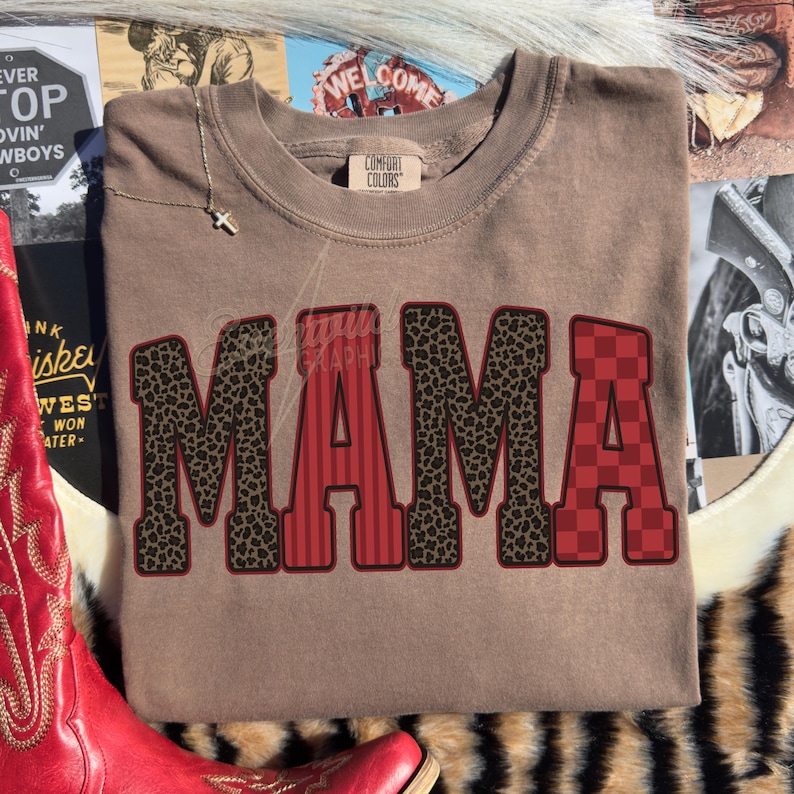 May include: A tan short-sleeve t-shirt with a crew neckline featuring the word MAMA printed in large block letters. The letters alternate between a brown leopard print pattern and a red pattern with vertical stripes or a checkered design.