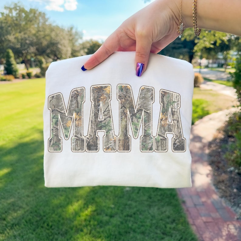 May include: White sweatshirt with the word "MAMA" in large, arched letters. The letters are filled with a camouflage pattern. The sweatshirt is held up against a background of green grass and trees.