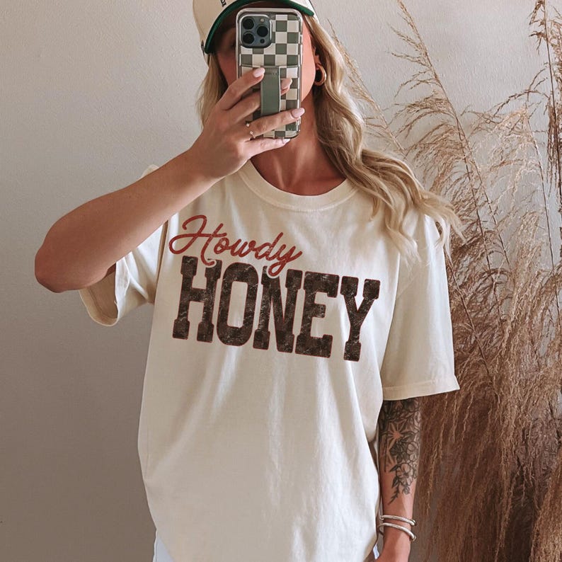 May include: Cream-colored t-shirt with the words "Howdy HONEY" in a vintage-style font. The shirt has short sleeves and a crew neck. The text is in a combination of red and brown colors. The shirt is part of a casual wear collection.
