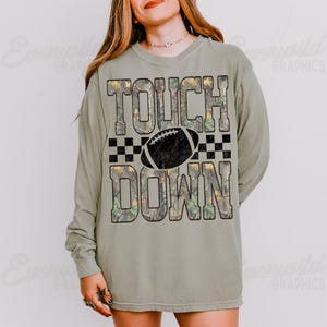 May include: A sage green long-sleeve t-shirt featuring the text TOUCH DOWN in a camouflage pattern with a black football graphic and a black checkered border. The design is centered on the chest of the shirt, which has a crew neckline.