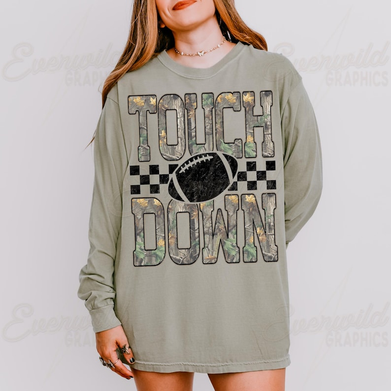 May include: A sage green long-sleeve t-shirt featuring the text TOUCH DOWN in a camouflage pattern with a black football graphic and a black checkered border. The design is centered on the chest of the shirt, which has a crew neckline.