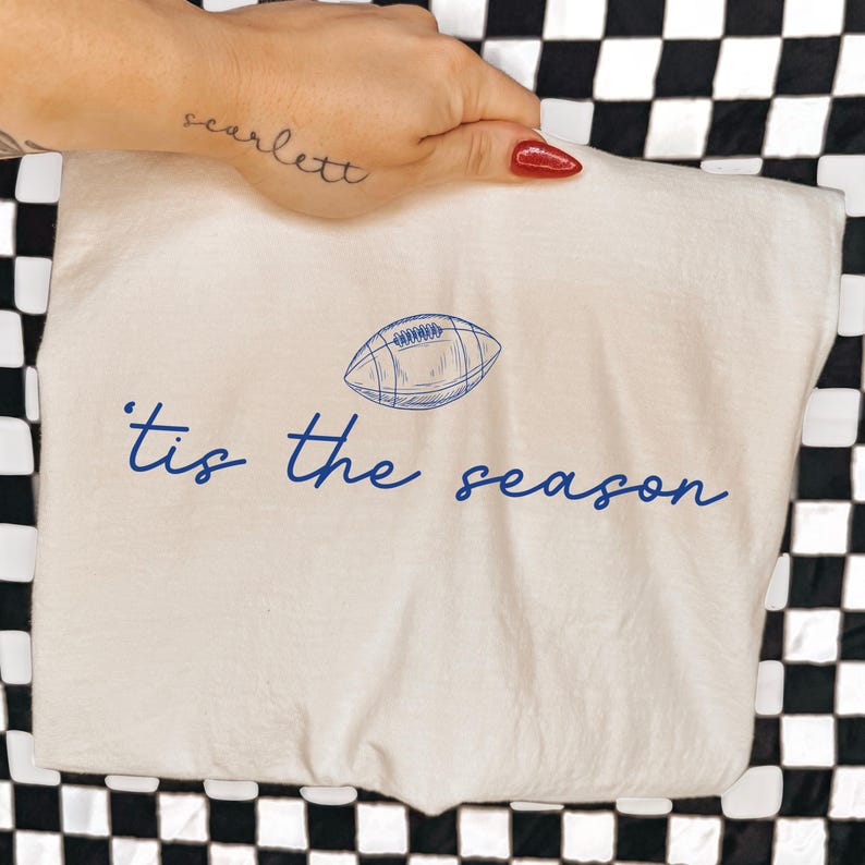 May include: A cream-colored t-shirt featuring a blue line-art illustration of an American football centered above the blue cursive text that reads 'tis the season'. The design is printed on the front of the garment in a simple, clean style.