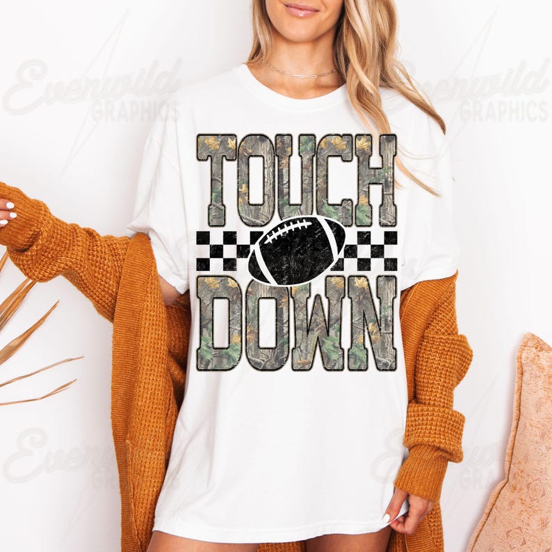 May include: A white short-sleeve t-shirt featuring the text TOUCH DOWN in a camouflage pattern. Between the words is a black and white checkered stripe and a black football graphic. The design is centered on the front of the garment.