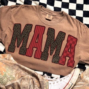 May include: A tan short-sleeve t-shirt featuring the word MAMA in large block letters. The letters M and M have a brown leopard print, while the letters A and A feature red vertical stripes and a red checkered pattern respectively.