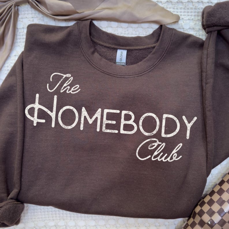 K&ouml;nnte beinhalten: Ein braunes Sweatshirt mit dem wei&szlig;en Schriftzug "The Homebody Club" in kursiver Schrift. Das Sweatshirt mit Rundhalsausschnitt ist aus weichem Material. Das Bild ist aus der Vogelperspektive aufgenommen.