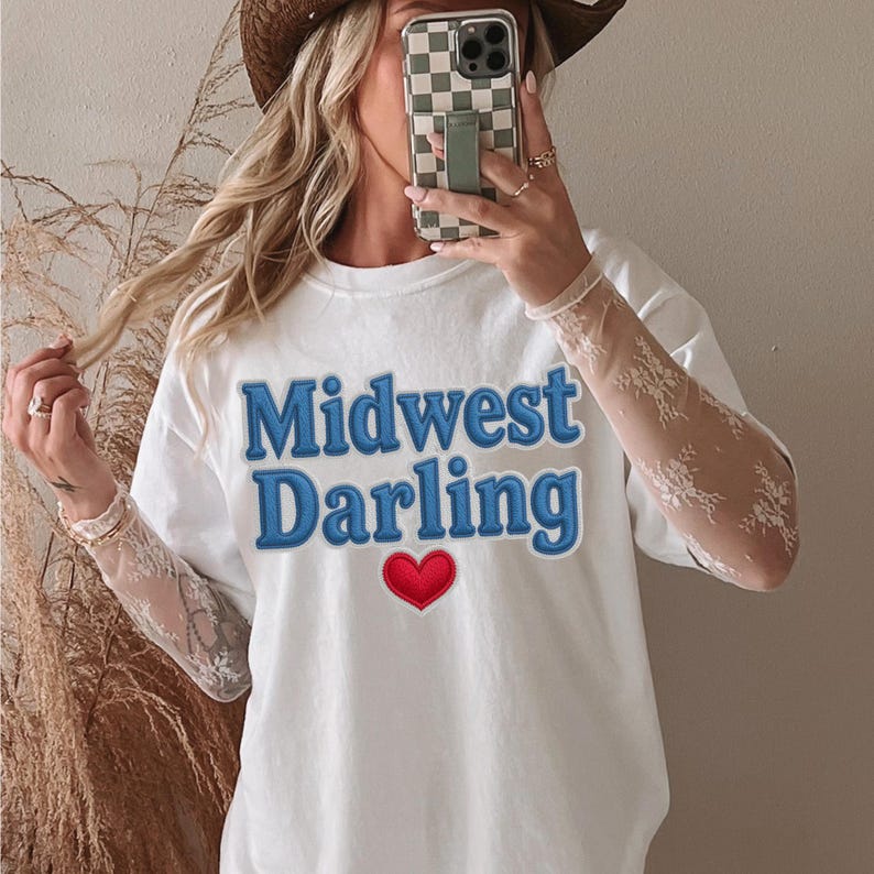 May include: White t-shirt with the text "Midwest Darling" in blue, outlined in a darker blue, and a red heart. The shirt is a casual style, suitable for everyday wear.