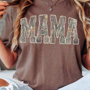 May include: A brown t-shirt with the word "MAMA" in large, block letters across the front. The letters are filled with a camouflage pattern and outlined in white. The shirt is a solid color and has a crew neck.