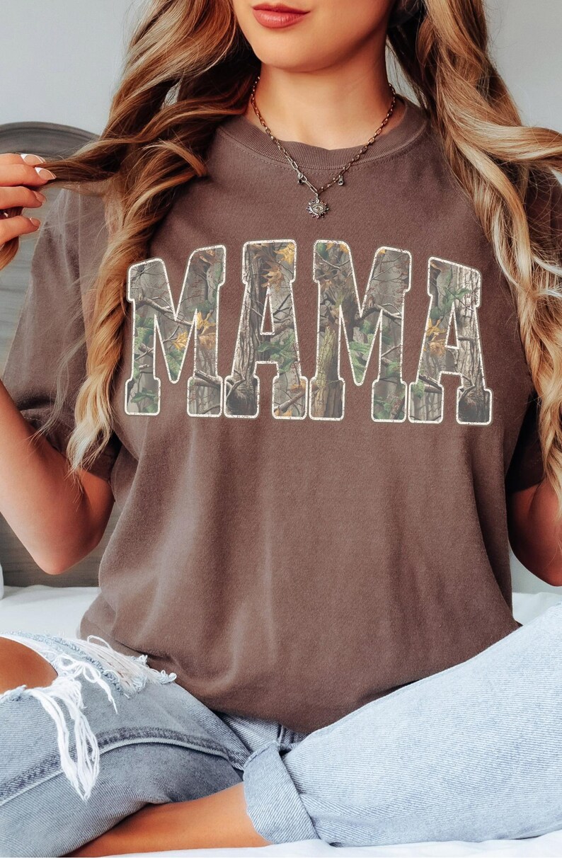 May include: A brown t-shirt with the word "MAMA" in large, block letters across the front. The letters are filled with a camouflage pattern and outlined in white. The shirt is a solid color and has a crew neck.