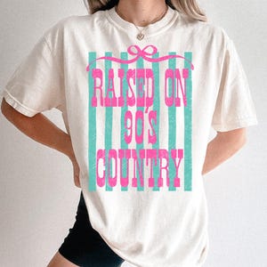 May include: Cream-colored t-shirt with the text "RAISED ON 90'S COUNTRY" in pink, set against a backdrop of vertical teal stripes. The design includes a pink bow at the top. The shirt is a casual, oversized fit.