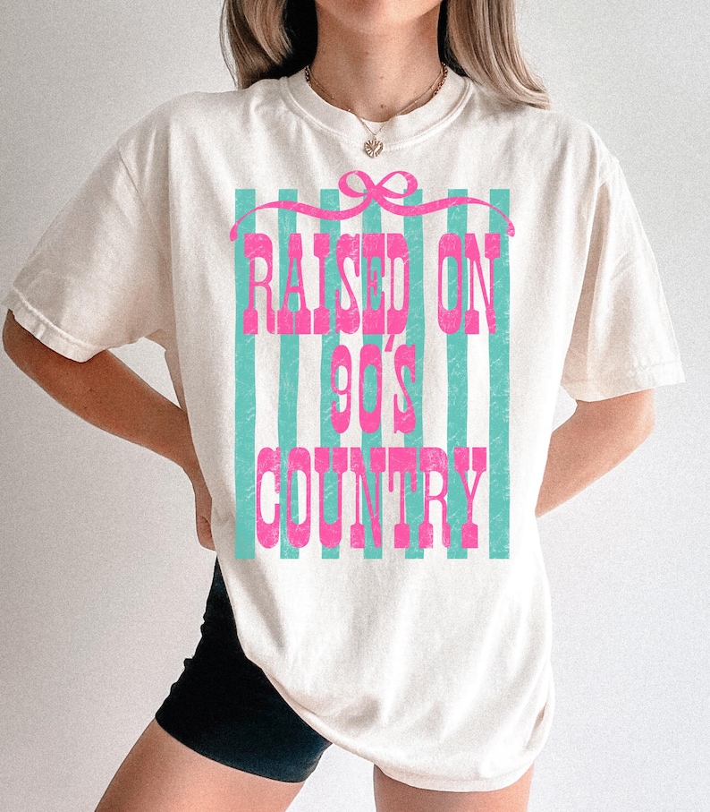 May include: Cream-colored t-shirt with the text "RAISED ON 90'S COUNTRY" in pink, set against a backdrop of vertical teal stripes. The design includes a pink bow at the top. The shirt is a casual, oversized fit.