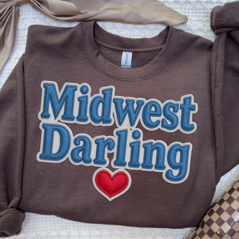 May include: A brown sweatshirt with the words "Midwest Darling" in blue, outlined in white, and a red heart. The sweatshirt is made of a soft material and is suitable for casual wear.