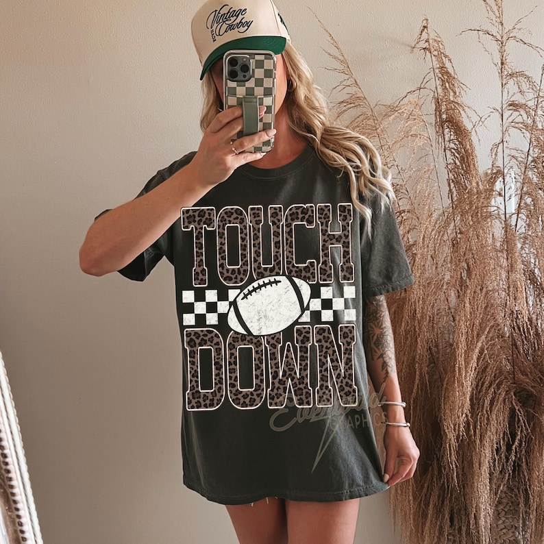 Puede incluir: Una camiseta de manga corta gris oscuro con el texto TOUCH DOWN en estampado de leopardo. Un gr&aacute;fico de bal&oacute;n de f&uacute;tbol americano blanco est&aacute; centrado entre las palabras, sobre un fondo de cuadros blanco y negro. Corte holgado.