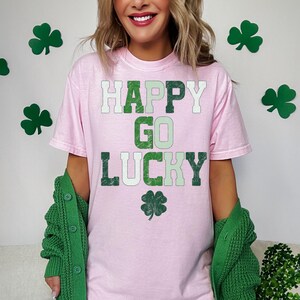 May include: A light pink t-shirt with the words "HAPPY GO LUCKY" in green and white lettering, along with a green shamrock. A green cardigan is draped over the shoulders. Green shamrock decorations are in the background.