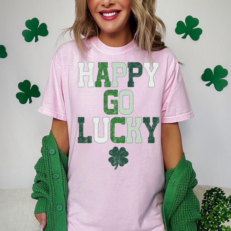 May include: A light pink t-shirt with the words "HAPPY GO LUCKY" in green and white lettering, along with a green shamrock. A green cardigan is draped over the shoulders. Green shamrock decorations are in the background.