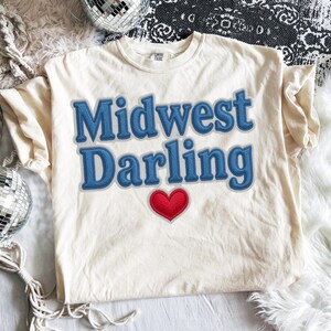 May include: Cream-colored t-shirt with the words "Midwest Darling" in blue, stitched-style lettering. A red heart is below the text. The shirt is on a white surface with disco balls.