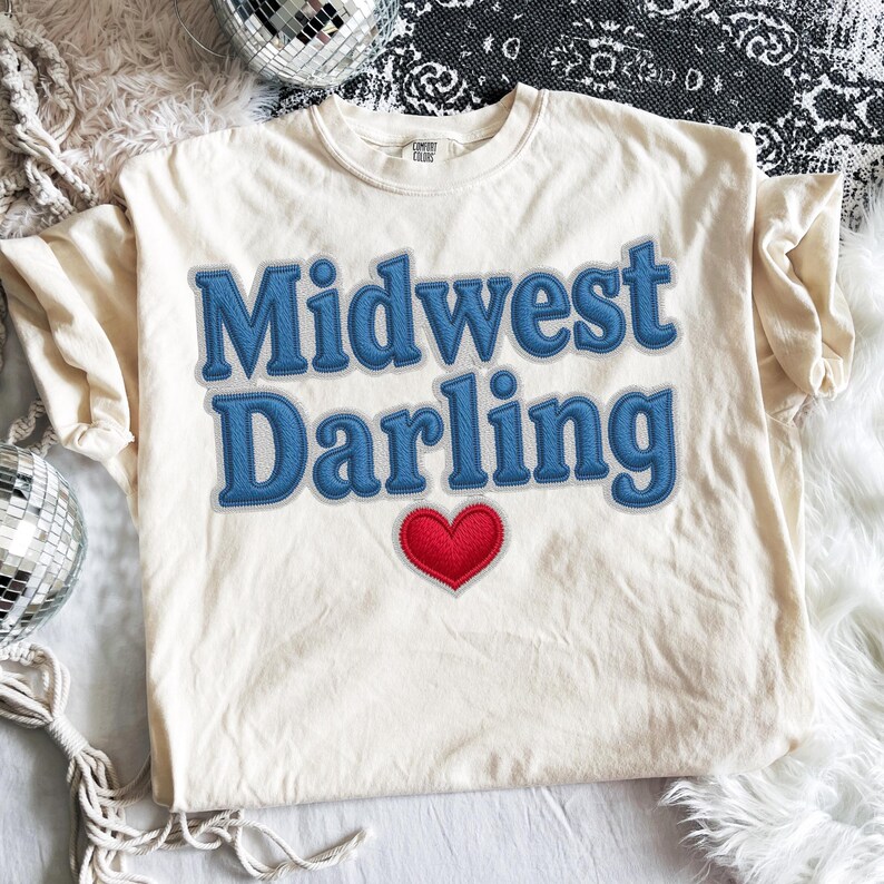 May include: Cream-colored t-shirt with the words "Midwest Darling" in blue, stitched-style lettering. A red heart is below the text. The shirt is on a white surface with disco balls.