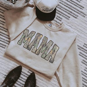 May include: A beige sweatshirt with the word "MAMA" in a camouflage design. A white and black trucker hat with the text "Howdy Cowboy" is above the sweatshirt. Black cowboy boots are at the bottom.