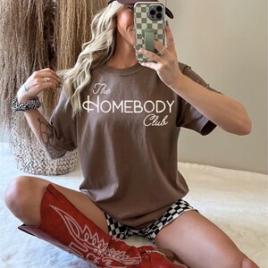 K&ouml;nnte beinhalten: Ein braunes T-Shirt mit dem Text "The Homebody Club" in wei&szlig;er Schrift. Die Person tr&auml;gt eine braune und beige Trucker-M&uuml;tze, rote Cowboy-Stiefel und schwarz-wei&szlig; karierte Shorts. Ein Telefon mit einer gr&uuml;n karierten H&uuml;lle befindet sich vor dem Gesicht der Person.