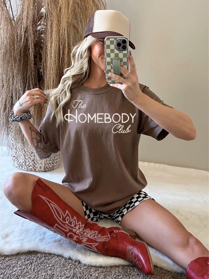 K&ouml;nnte beinhalten: Ein braunes T-Shirt mit dem Text "The Homebody Club" in wei&szlig;er Schrift. Die Person tr&auml;gt eine braune und beige Trucker-M&uuml;tze, rote Cowboy-Stiefel und schwarz-wei&szlig; karierte Shorts. Ein Telefon mit einer gr&uuml;n karierten H&uuml;lle befindet sich vor dem Gesicht der Person.