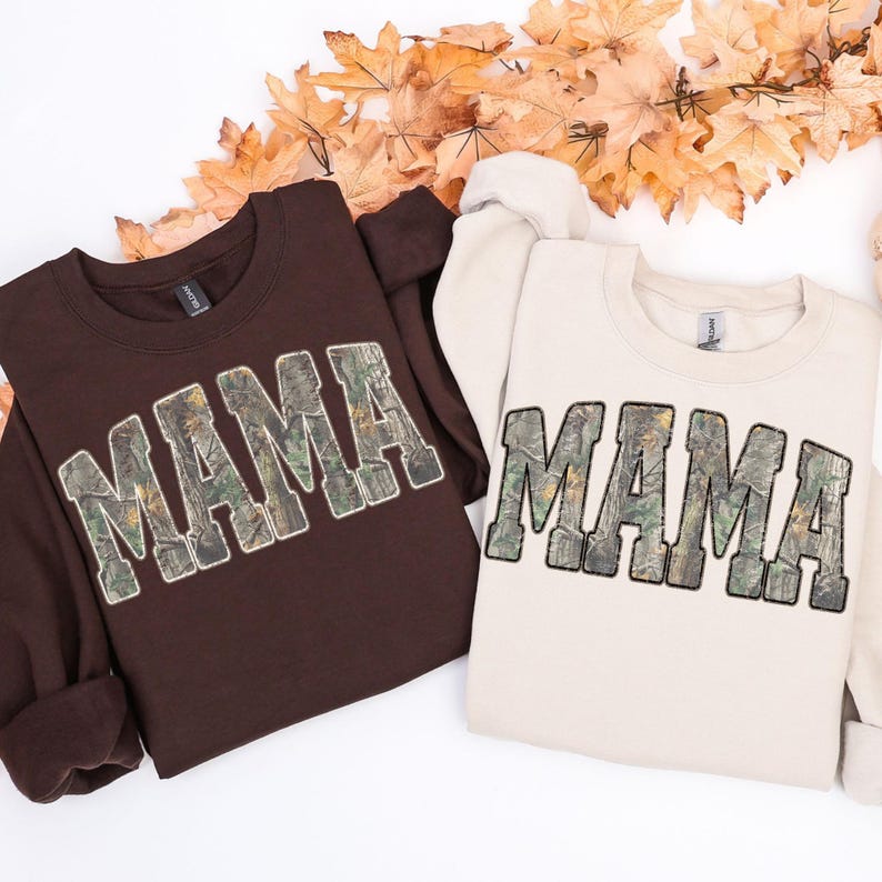 May include: Two sweatshirts, one brown and one cream, each with the word "MAMA" in a camouflage pattern across the front. The sweatshirts are laid on a white surface with autumn leaves.