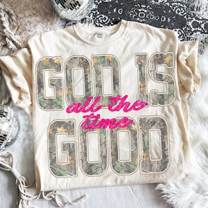 K&ouml;nnte beinhalten: Cremefarbenes T-Shirt mit dem Text "GOD IS GOOD all the time". Die Buchstaben "GOD IS GOOD" sind in einem Camouflage-Muster gehalten, "all the time" in Pink. Rundhalsausschnitt.