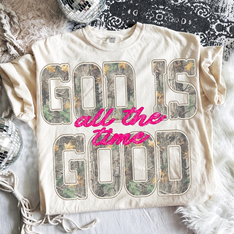 K&ouml;nnte beinhalten: Cremefarbenes T-Shirt mit dem Text "GOD IS GOOD all the time". Die Buchstaben "GOD IS GOOD" sind in einem Camouflage-Muster gehalten, "all the time" in Pink. Rundhalsausschnitt.