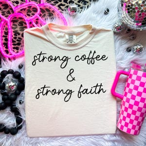 May include: Cream-colored t-shirt with the words "strong coffee & strong faith" in black script. A pink checkered tumbler with a handle is on the right. The shirt is folded and displayed on a white fluffy surface.
