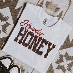 May include: White t-shirt with the words "Howdy HONEY" in a distressed, maroon and brown font. The shirt is laid on a patterned surface, with a pair of white and brown sneakers visible in the lower left corner.