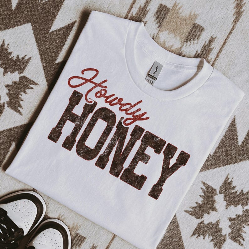 May include: White t-shirt with the words "Howdy HONEY" in a distressed, maroon and brown font. The shirt is laid on a patterned surface, with a pair of white and brown sneakers visible in the lower left corner.