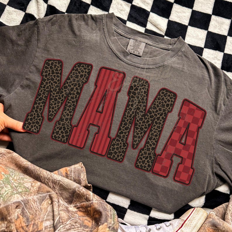 May include: A charcoal gray short-sleeve t-shirt featuring the word MAMA in large block letters. The letters alternate between a black and brown leopard print, a red and dark red vertical stripe pattern, and a red and dark red checkered pattern.