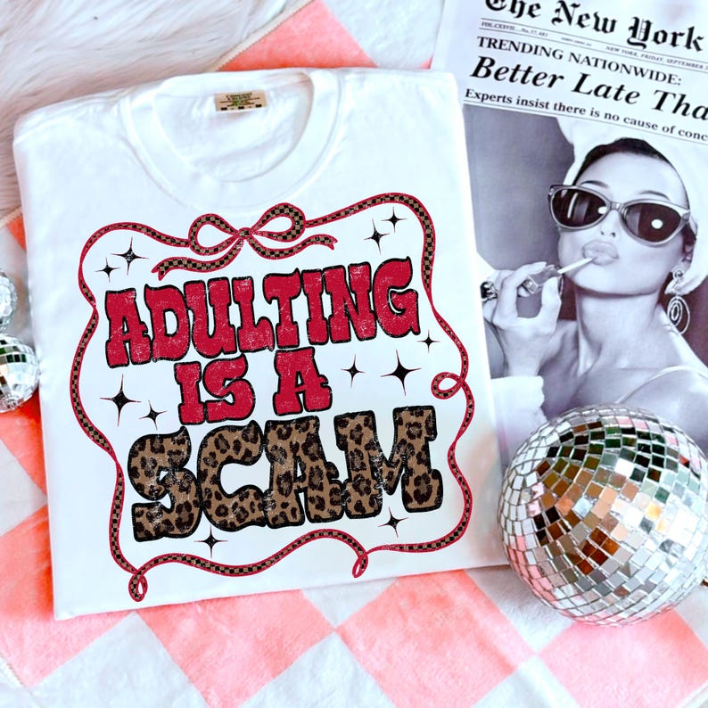 May include: White t-shirt featuring the text "ADULTING IS A SCAM" in red and leopard print. The design is framed with a red and black border. The shirt is placed on a pink and white checked surface.