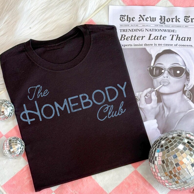 K&ouml;nnte beinhalten: Schwarzes T-Shirt mit Rundhalsausschnitt und dem Schriftzug "The Homebody Club" in hellblauer Schrift. Das Shirt wird mit einer Zeitung und Discokugeln pr&auml;sentiert.
