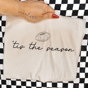 May include: A cream-colored t-shirt featuring a black line-art illustration of an American football centered above the black cursive text that reads 'tis the season'. The design is printed on the front of the fabric with a simple, clean aesthetic.