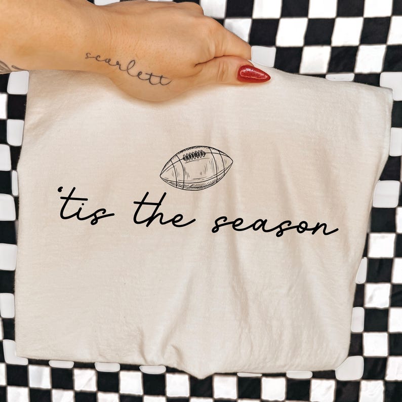 May include: A cream-colored t-shirt featuring a black line-art illustration of an American football centered above the black cursive text that reads 'tis the season'. The design is printed on the front of the fabric with a simple, clean aesthetic.