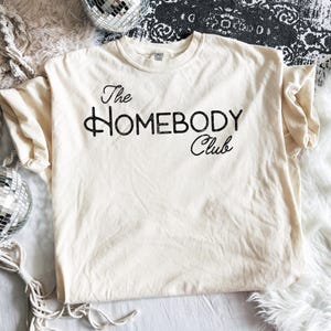 K&ouml;nnte beinhalten: Cremefarbenes T-Shirt mit dem Text "The Homebody Club" in schwarzer Schrift. Das Shirt ist aus einem weichen Material und hat einen klassischen Rundhalsausschnitt. Das Shirt liegt flach auf einer wei&szlig;en Oberfl&auml;che.