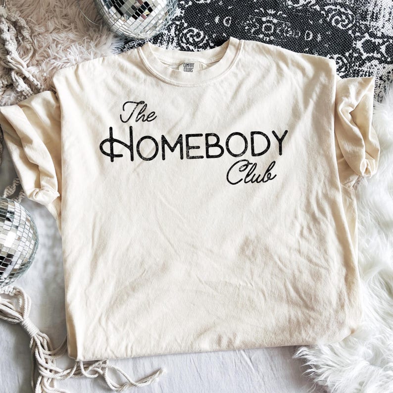 K&ouml;nnte beinhalten: Cremefarbenes T-Shirt mit dem Text "The Homebody Club" in schwarzer Schrift. Das Shirt ist aus einem weichen Material und hat einen klassischen Rundhalsausschnitt. Das Shirt liegt flach auf einer wei&szlig;en Oberfl&auml;che.