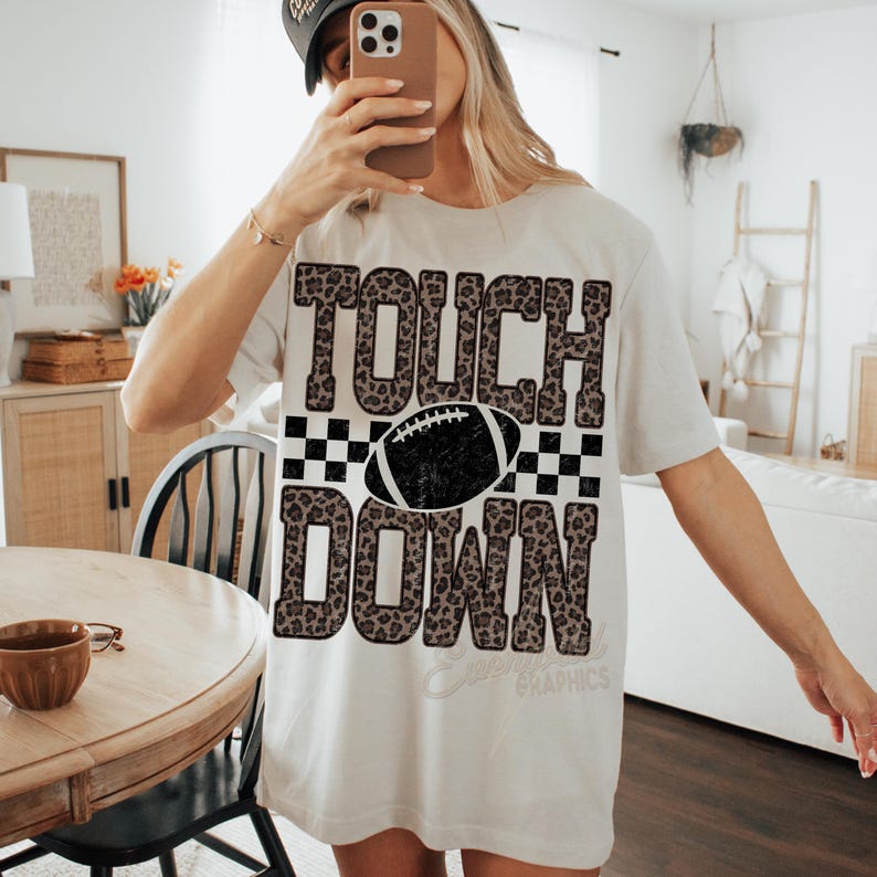 Puede incluir: Una camiseta de manga corta color beige claro con un estampado gr&aacute;fico que incluye el texto TOUCH DOWN en un patr&oacute;n de leopardo marr&oacute;n. Un borde a cuadros blanco y negro rodea una ilustraci&oacute;n de un bal&oacute;n de f&uacute;tbol americano en el centro.