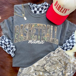 May include: A grey short-sleeved shirt with the words "BASKETBALL mama" in a camouflage design. A red and white baseball cap with the word "DARLIN'" is on top. A gold necklace and camouflage trousers are also visible.