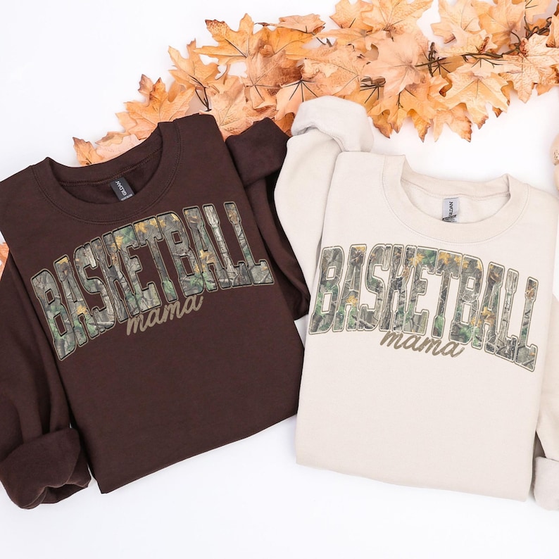 May include: Two sweatshirts, one brown and one cream, with the text "BASKETBALL mama" in a camouflage design. The sweatshirts are laid on a white surface with autumn leaves.