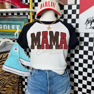May include: A white and black raglan t-shirt featuring the word MAMA in a mix of leopard print and red checkered patterns. The shirt is displayed on a mannequin wearing a white cap with the word DARLIN in red and a gold star pendant necklace.