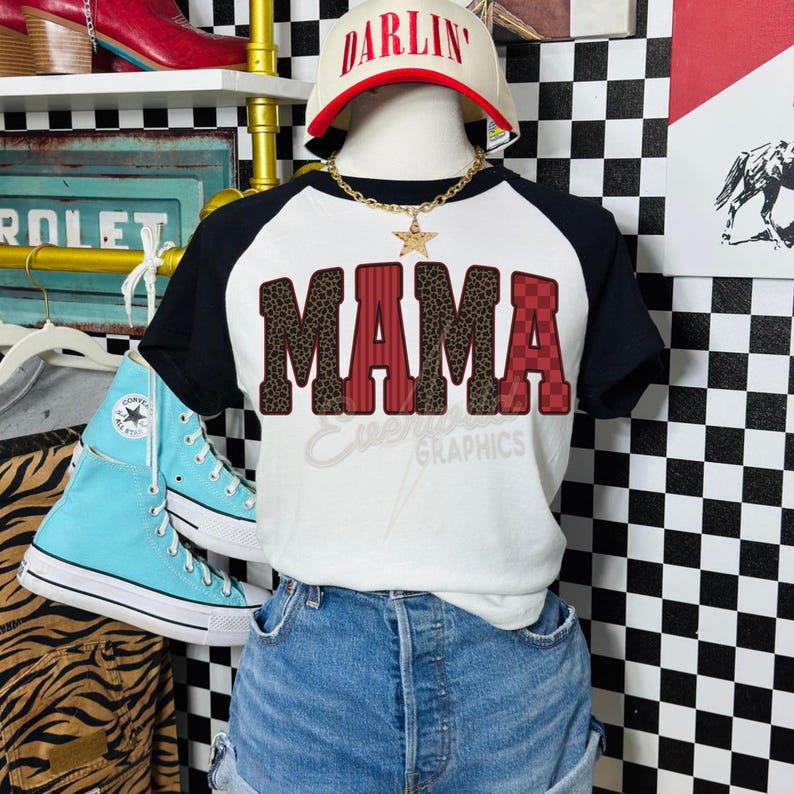 May include: A white and black raglan t-shirt featuring the word MAMA in a mix of leopard print and red checkered patterns. The shirt is displayed on a mannequin wearing a white cap with the word DARLIN in red and a gold star pendant necklace.