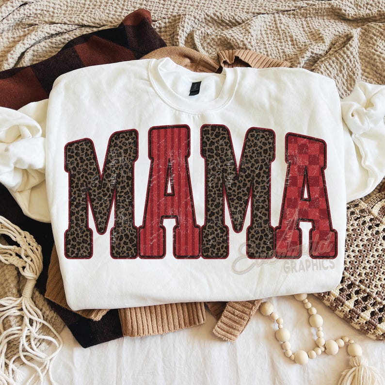 May include: A white crewneck sweatshirt featuring the word MAMA in large block letters. The letters M, M, and A have a brown and black leopard print pattern, while the letters A and A feature red vertical stripes and a red checkered pattern.
