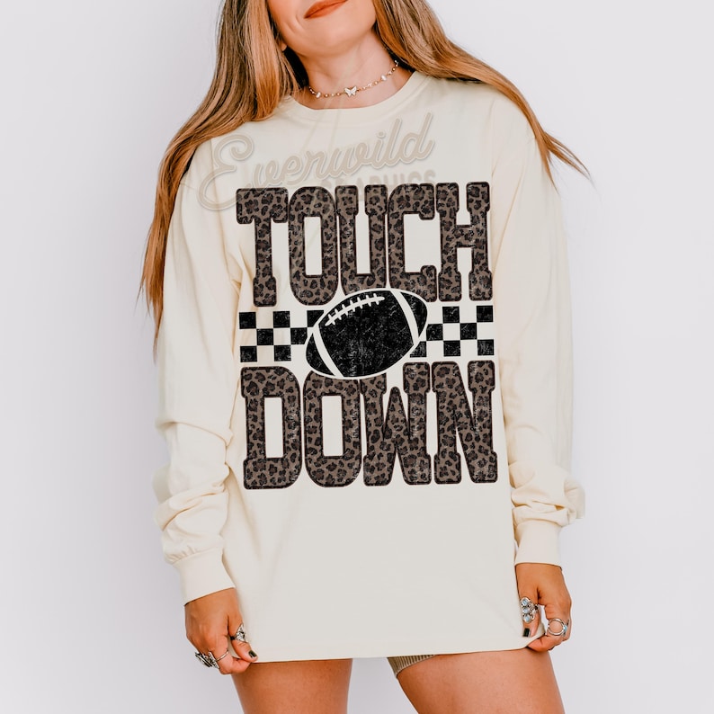 Puede incluir: Una camiseta de manga larga color crema con el texto TOUCH DOWN en un patr&oacute;n de leopardo marr&oacute;n. Una franja a cuadros blancos y negros separa las palabras, con un gr&aacute;fico de bal&oacute;n de f&uacute;tbol americano negro centrado. El dise&ntilde;o tiene textura desgastada.