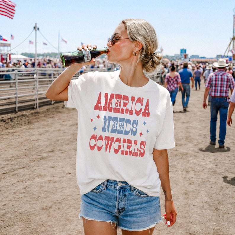 Może przedstawiać: Biała koszulka z napisem "America Needs Cowgirls" w czerwonych i niebieskich literach. Koszulka jest połączona z dżinsowymi szortami. Osoba pije z butelki Coca-Coli w tłumie.