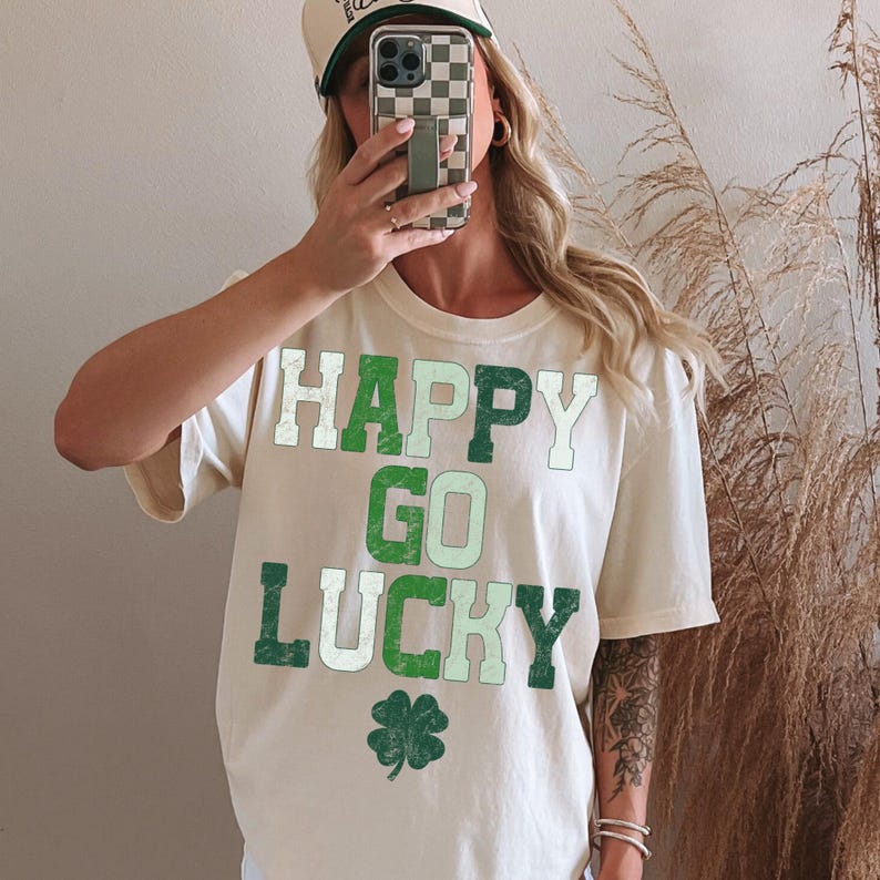 May include: Cream-colored t-shirt with the words "HAPPY GO LUCKY" in green and white block letters, along with a green shamrock. The person is wearing a baseball cap with a green brim.